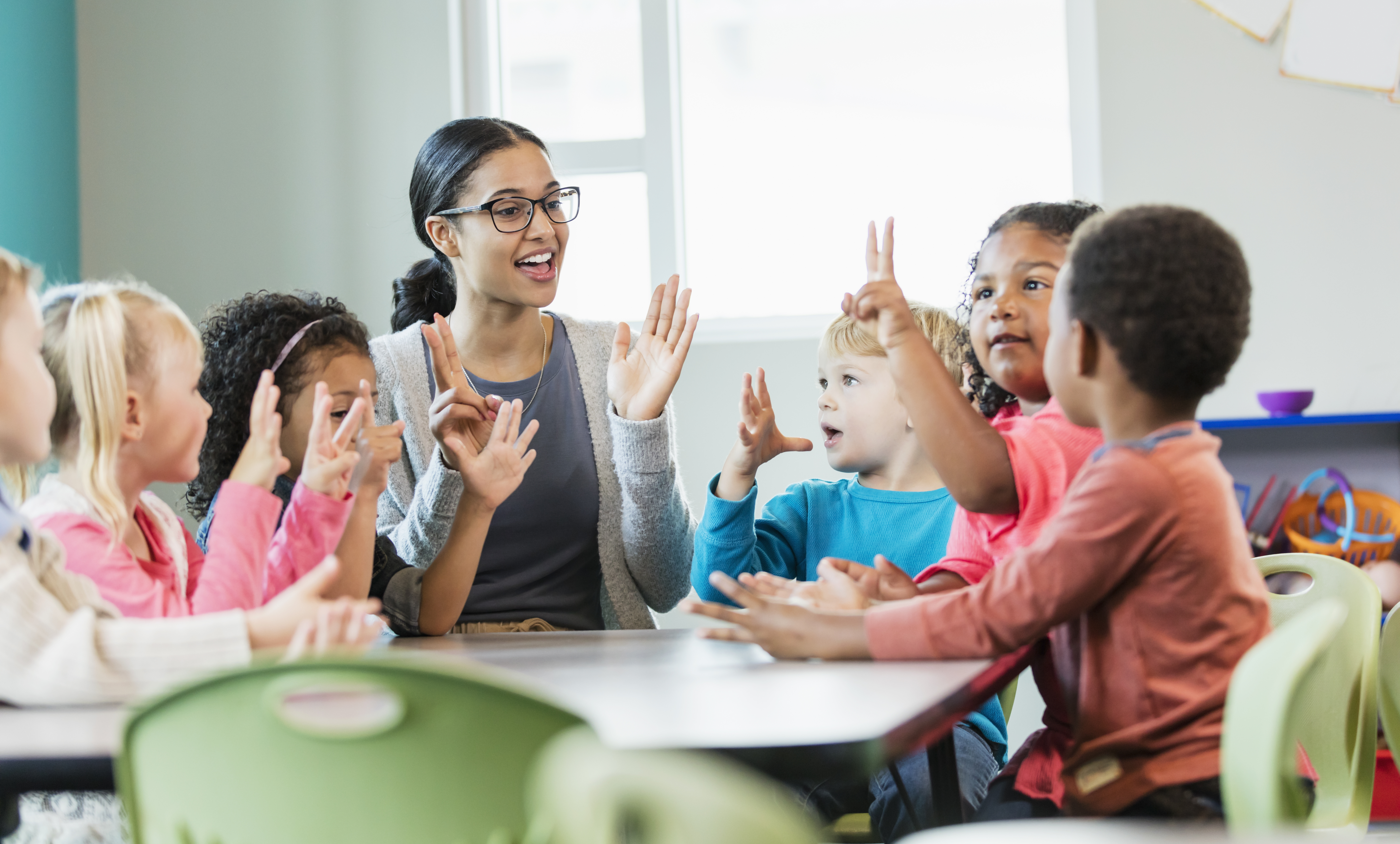 A multi-ethnic group of six preschool children with a mixed race African-American and Caucasian teacher, sitting around a table in a classroom. The teacher and some of her students have their hands raised, holding up fingers, learning how to count.